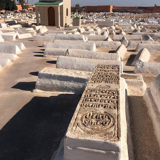 The Jewish cemetery of Marrakech — hundreds of white plaster tombs arranged in dense rows across a walled enclosure, their surfaces worn smooth by sun and time, with a small mosque-like structure visible at the far end under the deep blue Moroccan sky